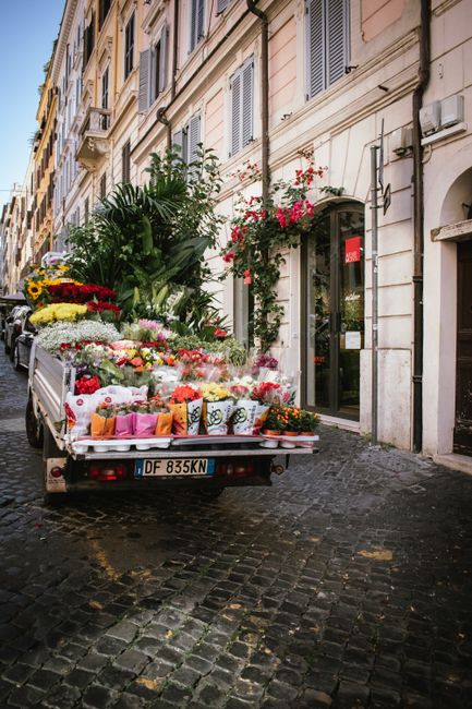 A delivery truck parked on a cobblestone street in front of a historic building with cream-colored walls and shuttered windows, loaded with a large array of fresh flowers and floral arrangements. The